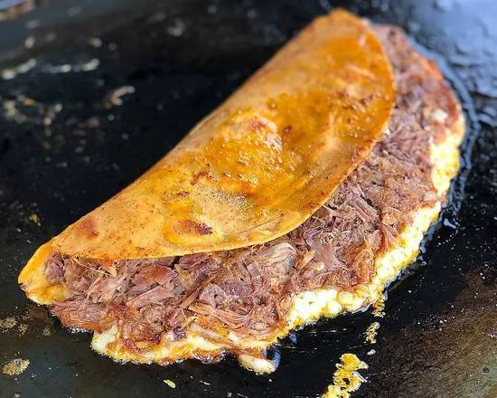 Quesabirria dorada sobre la plancha, con el queso derretido saliendo por el borde.