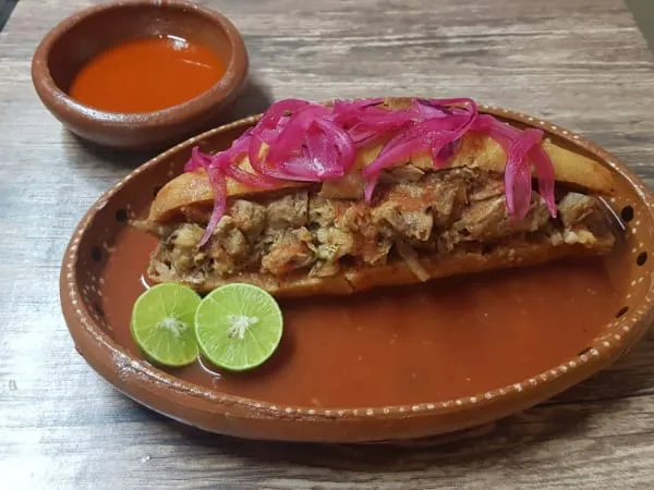 Torta ahogada de birria con cebolla morada, servida en plato de barro sobre salsa roja.